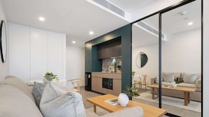 A modern living room with a sofa, coffee table, and a kitchen area with a dining set. - Modern Studio With Rooftop and BBQ Access (Perth)