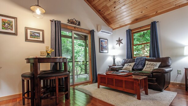 A cozy living room with a wooden ceiling, a leather sofa, a coffee table, and a dining area with a table and chairs. - Starry Night- Honeymoon Cabin| Hot Tub| Near GSMNP (Bryson City)