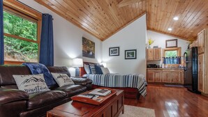 A cozy living room with a leather couch, a wooden coffee table, and a striped bedspread. There is a kitchen area with wooden cabinets and a refrigerator. - Starry Night- Honeymoon Cabin| Hot Tub| Near GSMNP (Bryson City)