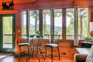 Una cabaña de madera con una mesa y sillas, vista a árboles y montañas, y una puerta que da al exterior.