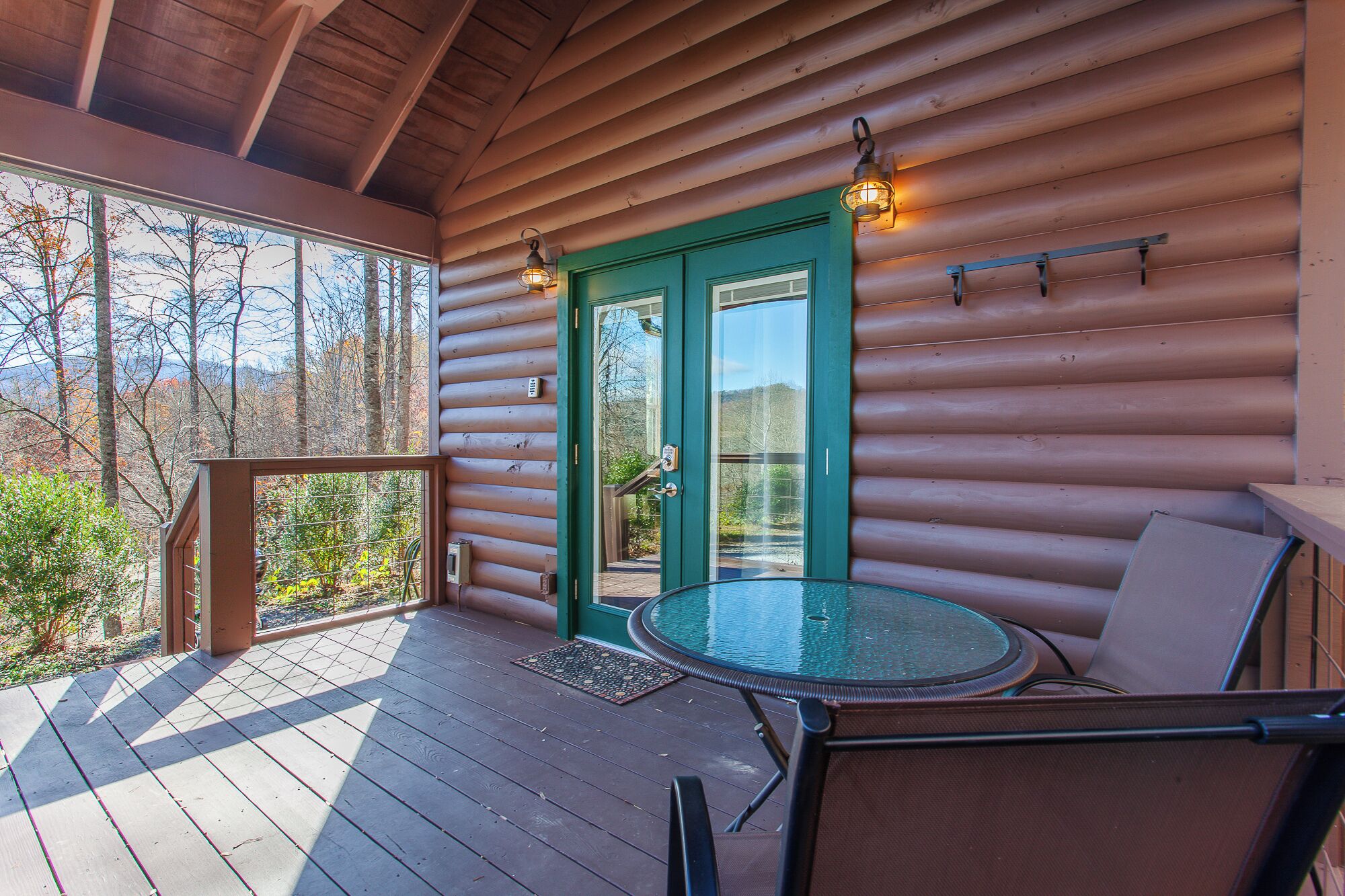Una cabaña de madera con puerta de cristal, una terraza con mesa y sillas, y vistas a árboles y montañas.