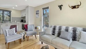 Een moderne woonkamer met een grijze bank, twee fauteuils, een houten salontafel, en een keuken op de achtergrond.