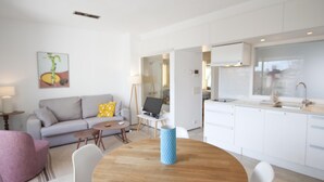 Un salon moderne comprenant un canapé, une table basse et une table à manger avec des chaises. On y trouve également un coin cuisine avec des meubles blancs et une fenêtre.