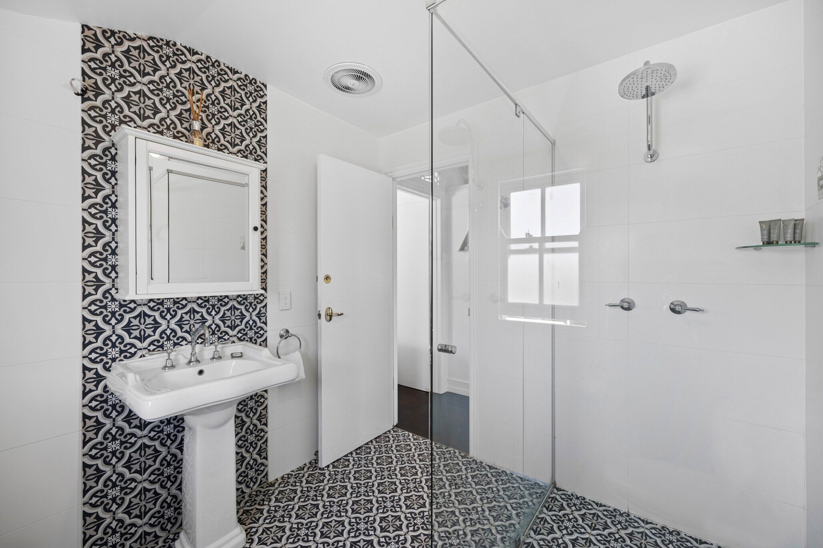 A bathroom with a patterned floor, a pedestal sink, a glass shower enclosure, and a white door.