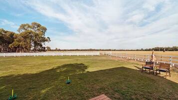 An outdoor equestrian arena with a wooden deck, white fences, and trees in the background.