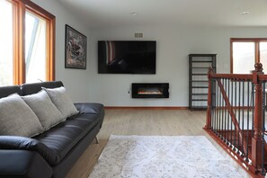 A living room with a black leather sofa, a fireplace, a TV mounted on the wall, and a wooden staircase.