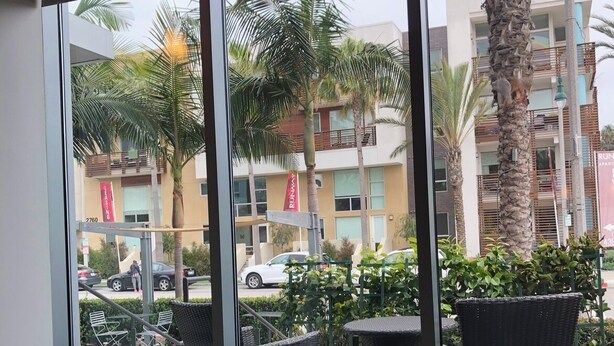 A hotel room with large windows, wicker furniture, and a view of palm trees and a building with balconies. - Cozy 1-bedroom condo loft with fitness room, AC in awesome LA/LAX/Beach (Los Angeles)