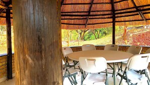 A round table with white chairs in a covered outdoor area with a thatched roof, surrounded by brick walls.