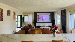 A spacious living room with a high ceiling, a flat-screen TV mounted on the wall, a dining area with a marble table, and a cozy seating arrangement.