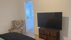 Makuuhuoneessa on sänky, puinen TV-taso, kuviollinen nojatuoli ja kylpyhuone, joka näkyy avoimen oven läpi.