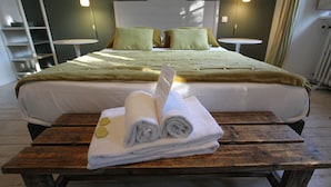 A neatly made bed with a green bedspread and two pillows, a wooden table with rolled towels, and a small round table in the background. - Chambre Tilleul 1 st floor (ST MALO)