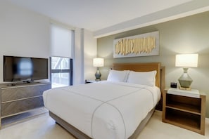 A modern hotel room with a large bed, a flat-screen TV, a desk with a lamp, and a wall-mounted artwork. - Sojourn | VA Square | 2 BR (Arlington)