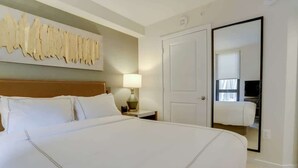 A hotel room with a large bed, a nightstand with a lamp, a wall-mounted artwork, a door, and a mirror reflecting part of the room. - Sojourn | VA Square | 1 BR (Arlington)