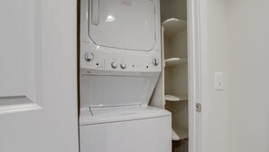 A compact laundry room with a front-loading washer and dryer, a control panel with multiple settings, and a small shelf above the dryer. - Sojourn | VA Square | 1 BR (Arlington)