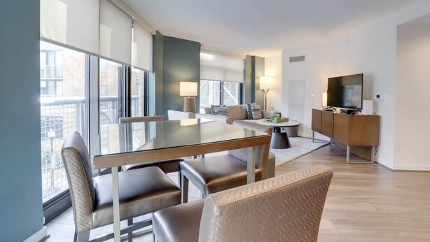 A modern living room with a glass dining table, comfortable seating, a sofa, a TV, and a cabinet. - Sojourn | VA Square | 1 BR (Arlington)
