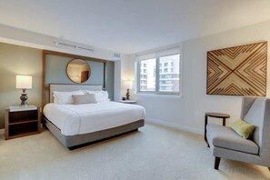 A modern hotel room with a large bed, bedside lamps, a chair, a window, and a decorative wall piece. - Sojourn | Randolph Towers (Arlington)