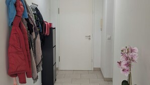 Käytävällä on kattoikkuna, takkien ripustamiseen tarkoitettu teline, jossa on useita takkeja, ruukkuorvidea ja musta metallipöytä.
