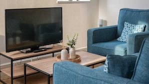 A modern living room with a flat-screen TV on a wooden coffee table, a blue sofa with patterned cushions, and two white vases with plants.
