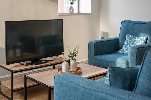 Un salon moderne avec un téléviseur à écran plat posé sur une table basse en bois, un canapé bleu agrémenté de coussins à motifs et deux vases blancs contenant des plantes.