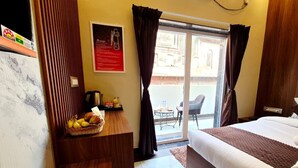A hotel room with a bed, a wooden nightstand with a fruit basket, a coffee machine, and a view of the outside through a glass door. - Ragam Residency (VARANASI)