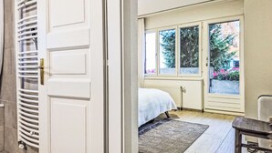Une chambre à coucher avec un lit, un radiateur, une fenêtre donnant sur des arbres et des fleurs, et un plancher en bois.