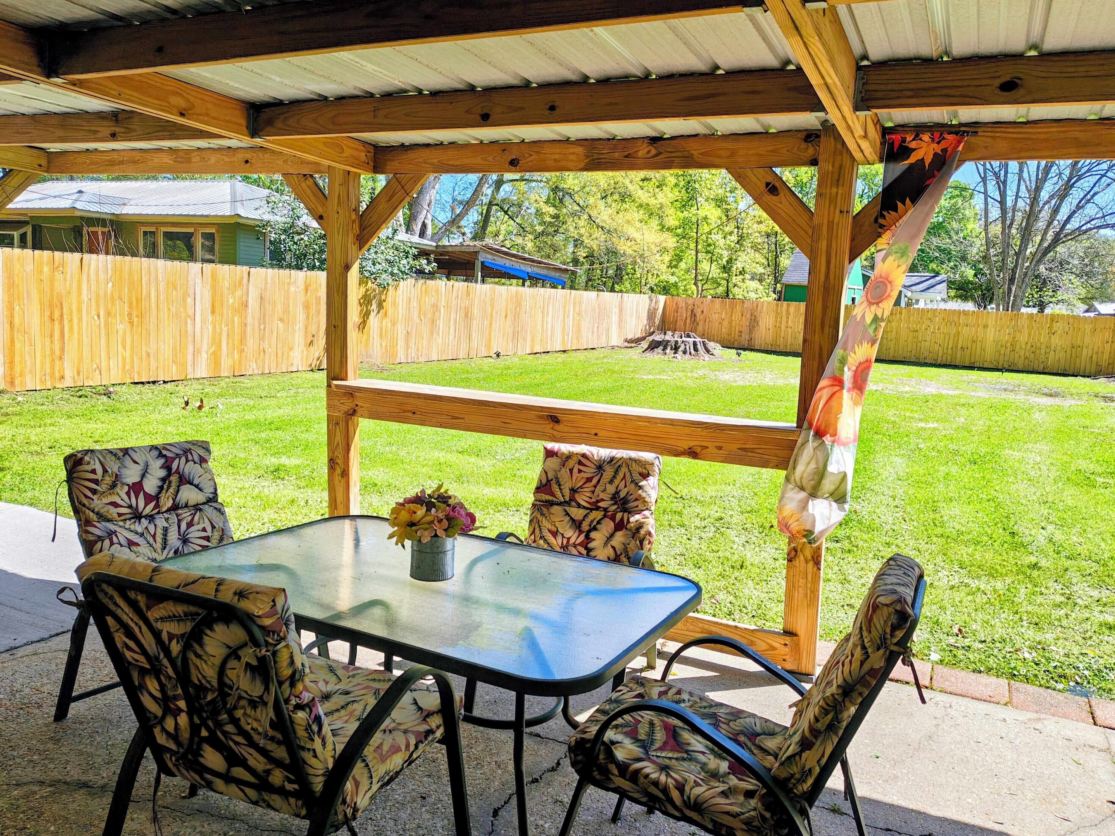 A covered patio with a table and chairs, a wooden fence, and a grassy yard.