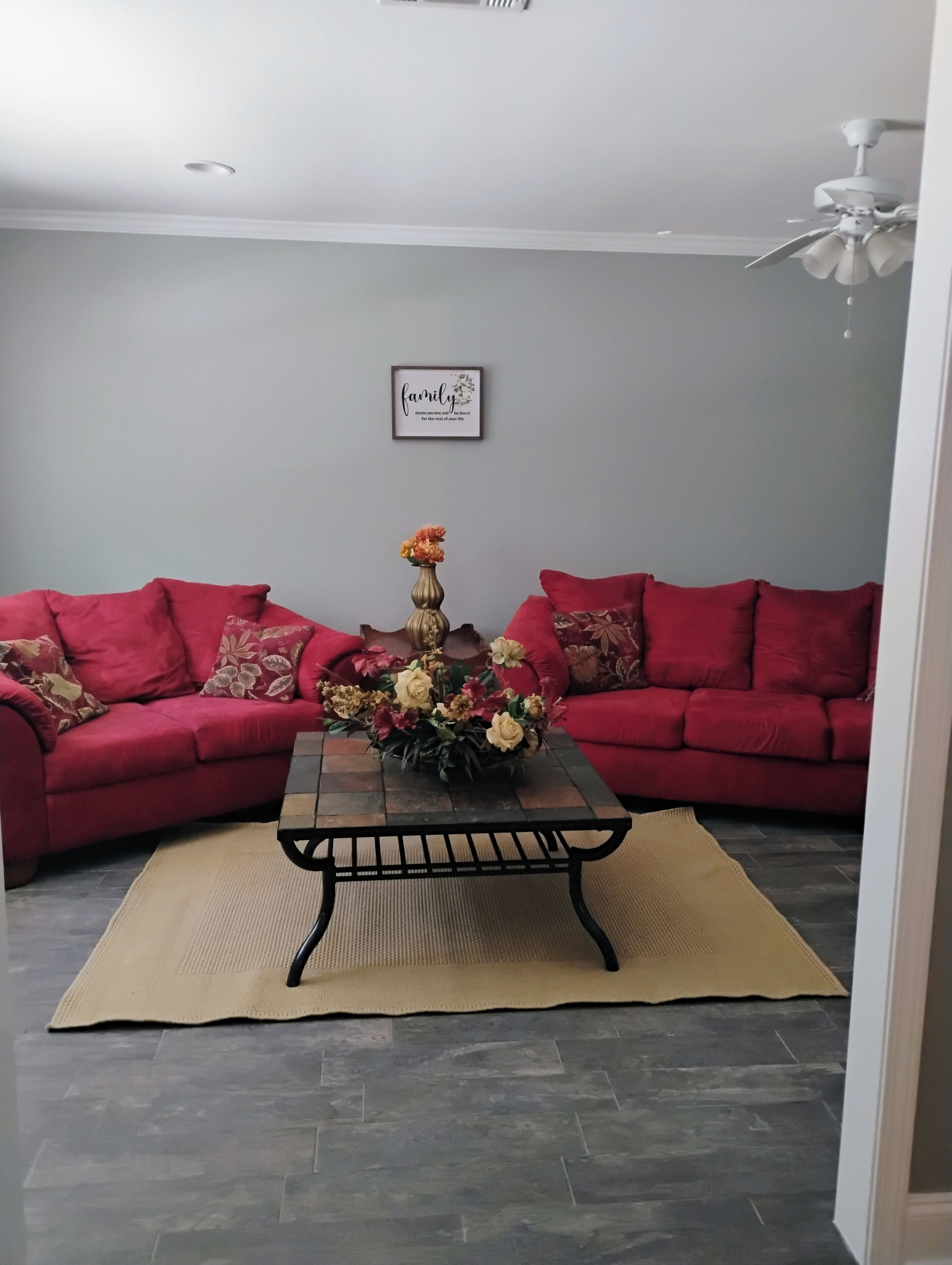 A living room with two red sofas, a coffee table with a floral centerpiece, a framed family quote on the wall, and a ceiling fan.