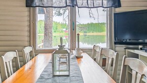 Un comedor con mesa de madera, sillas blancas, cortina negra, televisor y vistas a un lago y bosque.