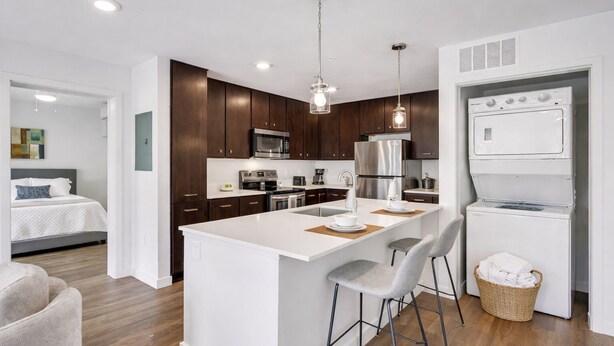 Una cocina moderna con alacenas de madera oscura, electrodomésticos de acero inoxidable, una isla blanca con taburetes y un área de lavandería con lavadora.