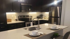 A modern kitchen with dark cabinetry, stainless steel appliances, and a central island with bar stools. - Luxury Austin Stay Near St. David’s with Pool+ Gym (Jarrell)