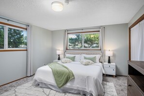 A bedroom with a large bed, two bedside tables with lamps, a window with curtains, and a view of trees outside. - 3/2 in Milwaukie - dead end street, free parking! (Milwaukie)