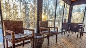 Une terrasse en bois avec des fauteuils en osier et une table, de grandes fenêtres donnant sur la forêt et un soleil éclatant.