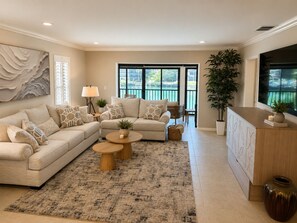 A living room with a sectional sofa, two coffee tables, a lamp, and a large window with a view of a pool. - Marco Island Waterfront Condo + Private Slip/Lift – Sea Ray Upgrade Option (Marco Island)