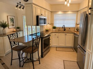 A kitchen with white cabinetry, a central island with bar stools, stainless steel appliances, and a window with blinds. - Marco Island Waterfront Condo + Private Slip/Lift – Sea Ray Upgrade Option (Marco Island)