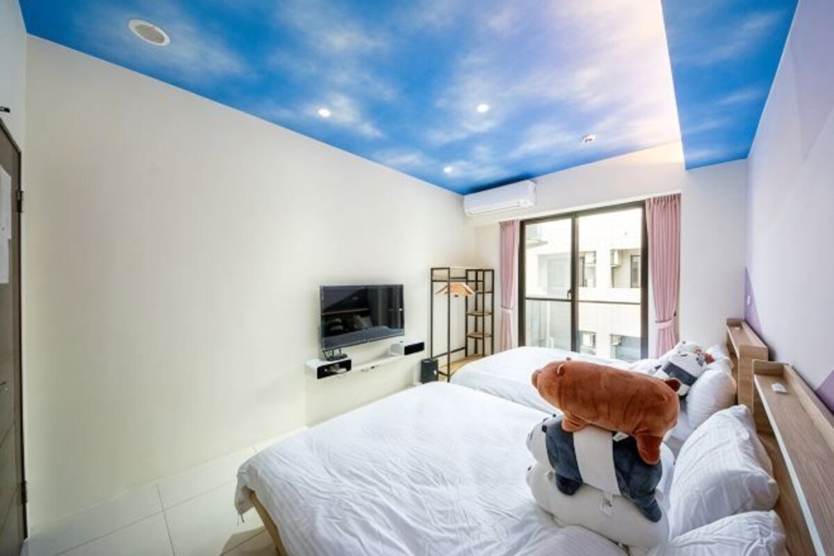 A hotel room with a bed, a television, a window, and a ceiling with a painted sky design.