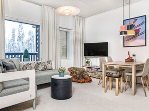 A modern living room with a sofa, a dining table with chairs, a TV, and a large window with a view of snow-covered trees. - Ruka chalets 3202 by Interhome (Kuusamo)