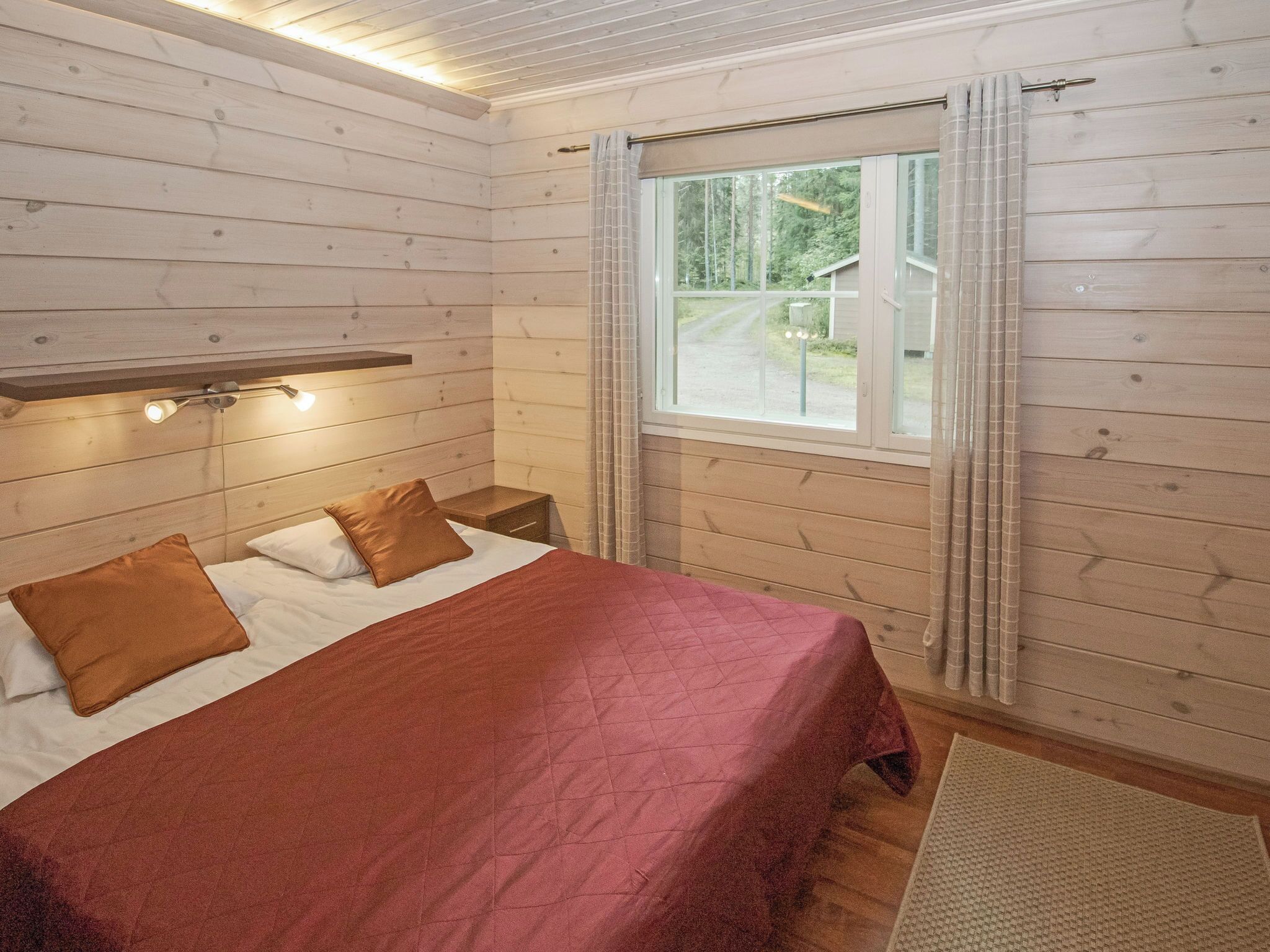 A wooden cabin bedroom with a bed, pillows, a nightstand, a window with curtains, and a shelf above the bed.
