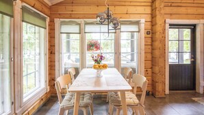 Interior de una cabaña de madera con una mesa de comedor, cuatro sillas, un jarrón de flores y ventanas amplias.