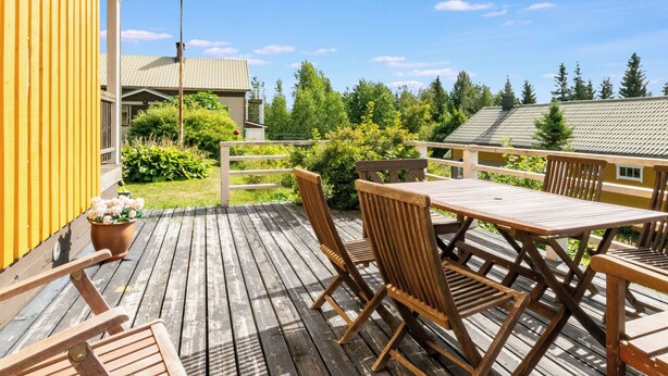 A wooden deck with a table and chairs, surrounded by a yellow building and greenery. - Vacation home Muhola by Interhome (Enonkoski)