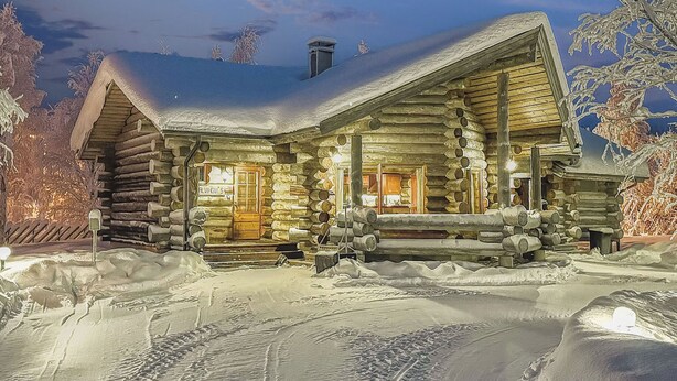 Eine Blockhütte mit schneebedecktem Dach, einer Veranda mit Feuerstelle und einem sichtbaren Schornstein.