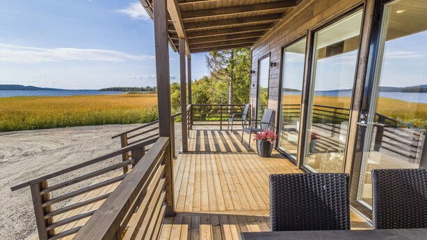 A wooden deck with a view of a lake and fields, featuring outdoor seating and a glass door leading to a covered area. - Niittyvilla by Interhome (Posio)