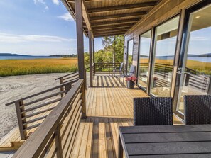 Una terraza de madera con vistas a un lago y campos, con mobiliario exterior y una puerta de cristal que da a una zona cubierta.