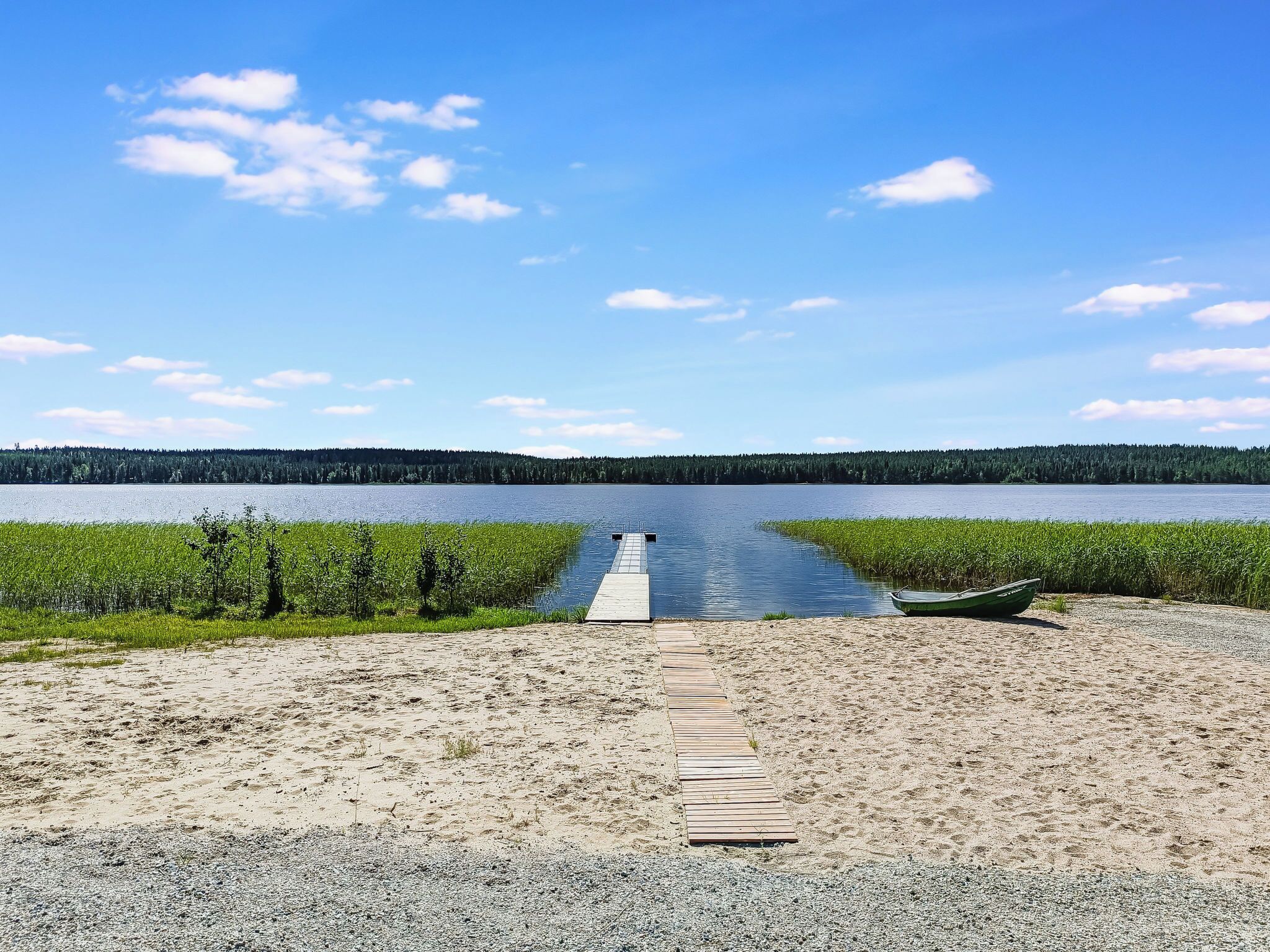 A gravel path leading to a wooden dock extending into a calm lake, surrounded by dense greenery and a clear sky.