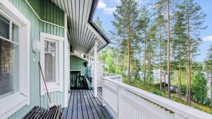 A wooden deck with a white railing, a green building with white trim, and a forested area in the background. - Saunaharju 6 by Interhome (Lieksa)