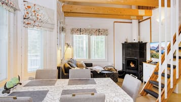 En hyggelig stue med pejs, et spiseområde med bord og stole, og en trappe til et øvre plan.