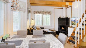 A cozy living room with a fireplace, a dining area with a table and chairs, and a staircase leading to an upper level. - Saunaharju 6 by Interhome (Lieksa)