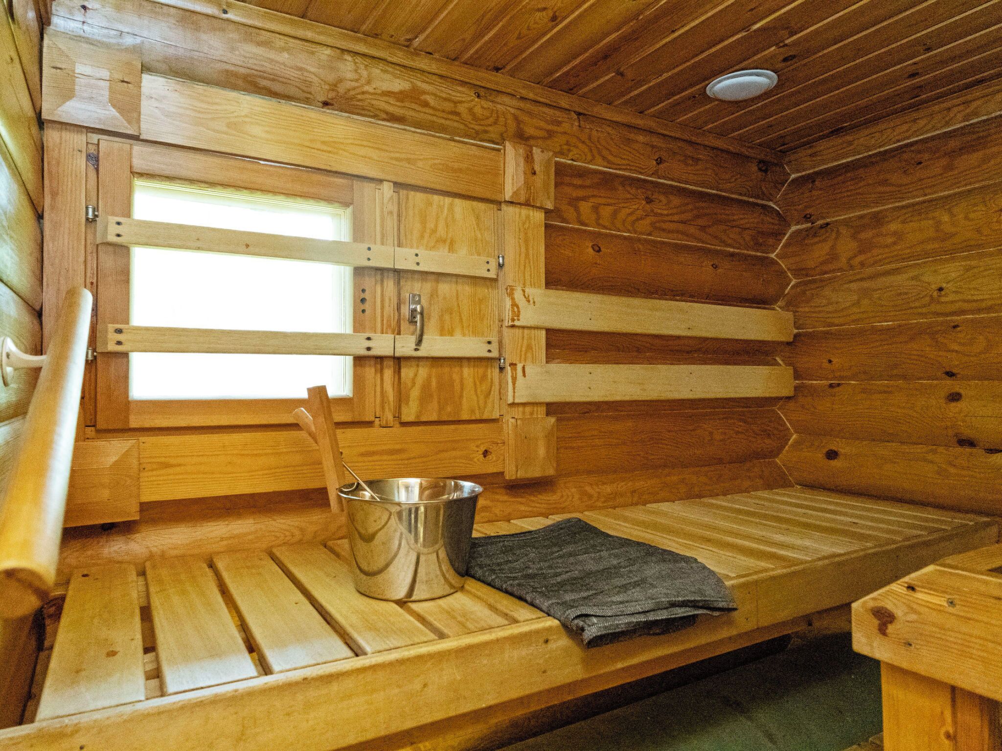 A wooden cabin interior with a bench, a metal bucket, and a window.