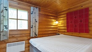 Une pièce de type cabane en bois, avec un lit, une fenêtre avec des rideaux, un radiateur mural et un panneau matelassé rouge.