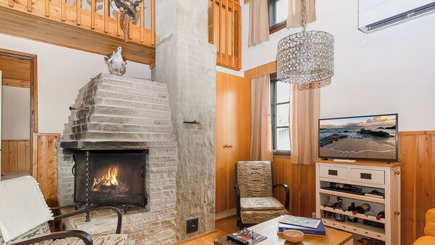Ein gemütliches Wohnzimmer mit Kamin aus Stein, Holzmöbeln, einem Fernseher und Blick ins Freie.