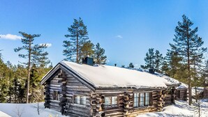 Une cabane en rondins dont le toit est recouvert de neige, entourée d’arbres et de neige.